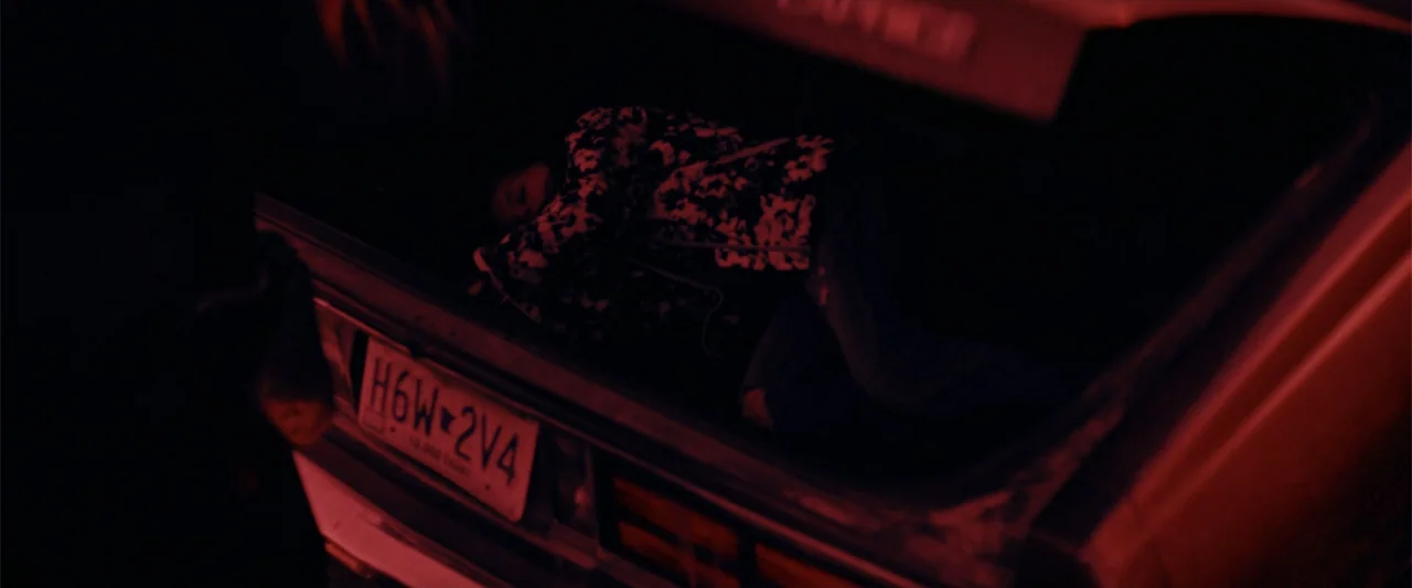 Silent Night, Deadly Night (2025) movie still: medium — An open trunk of a vehicle at night is illuminated by red light. A person reclines inside…