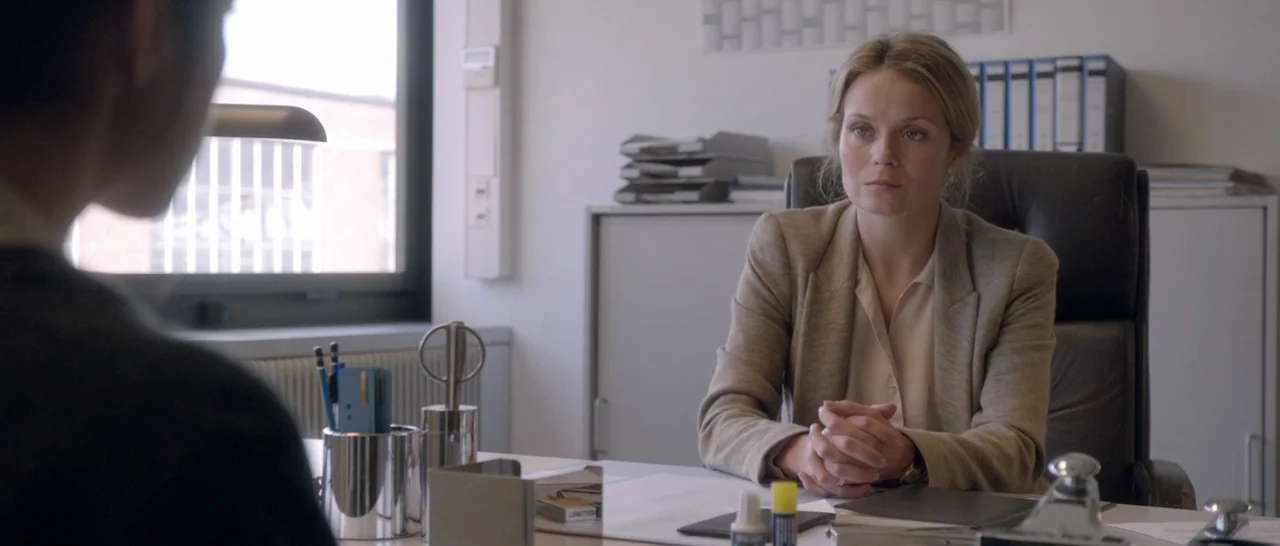 Nymphomaniac Vol 2 (2013) movie still: medium — An office interior features a white desk, filing cabinets, and a window with blinds. A blonde woman…