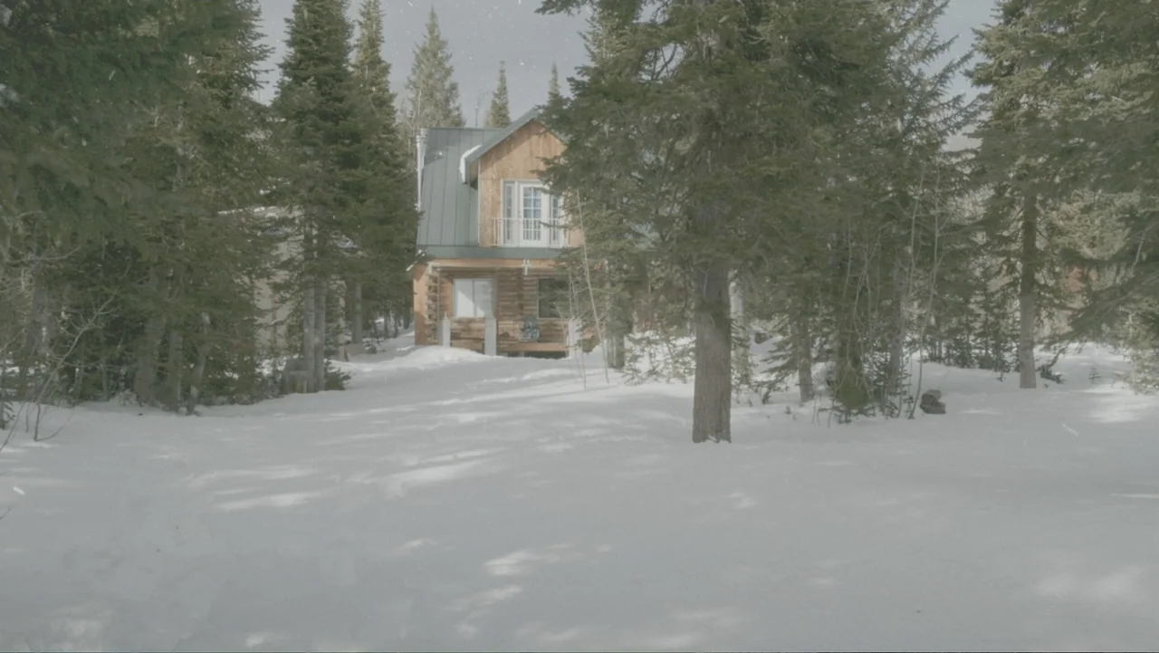 Alex & Emma (2003) movie still: establishing — A wooden cabin stands centered amidst a snowy clearing surrounded by dense pine trees. Falling snow…