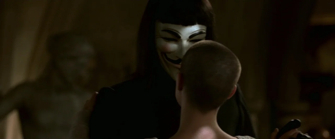 V for Vendetta (2006) movie still: medium — A dimly lit room contains stone walls and out-of-focus background details. A masked figure in a black…