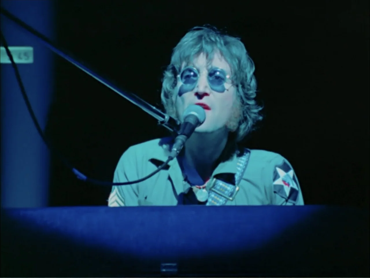 One to One: John & Yoko (2025) movie still: medium — A dark concert venue with the subject positioned behind a piano. A man in tinted glasses sings…