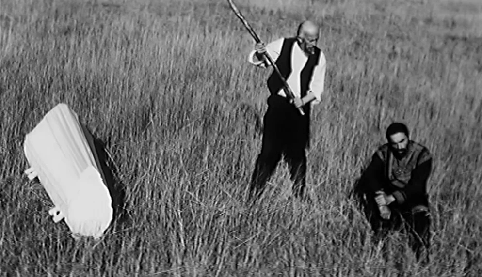 The Plea (1968) movie still: wide — A desolate field of tall grass sits under bright, harsh sunlight. An elderly man with a staff and a kneeling man…