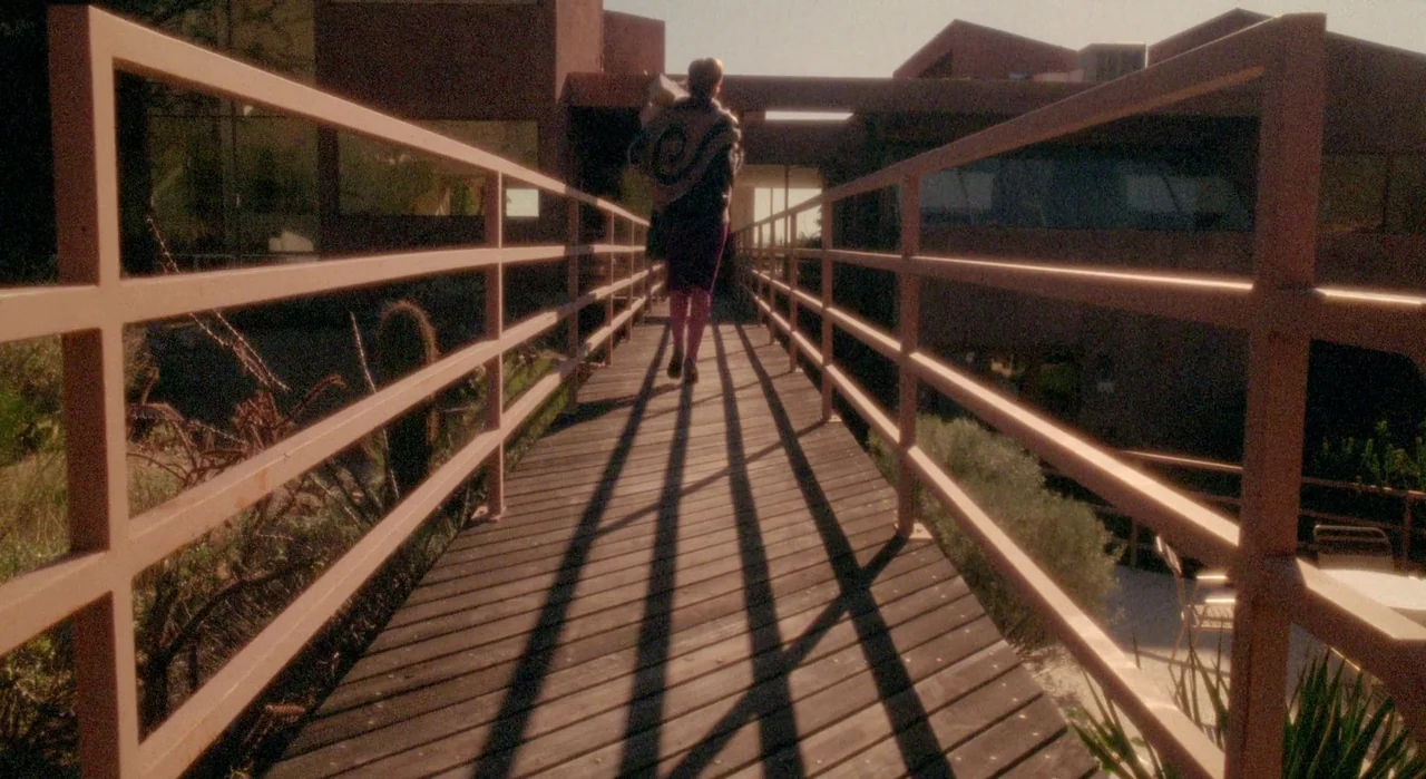 White Of The Eye (1987) movie still: wide — A wooden bridge connects two modern adobe-style buildings under bright sunlight. A person walks forward…