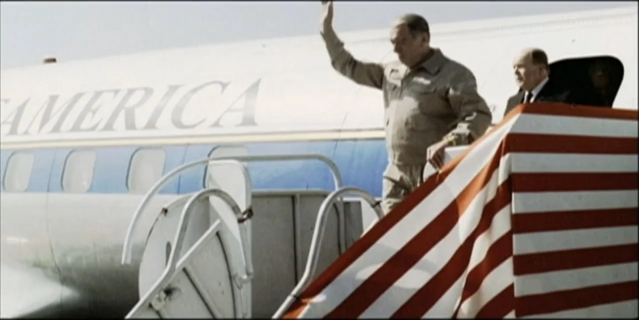 Path to War (2003) movie still: medium — An older man in a tan jacket stands on the mobile stairs of an airplane. He waves his hand while descending…