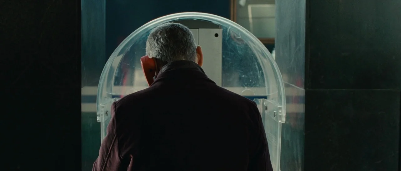 The American (2010) movie still: medium — An indoor public area featuring a clear plastic semi-circular phone booth structure. A man viewed from…