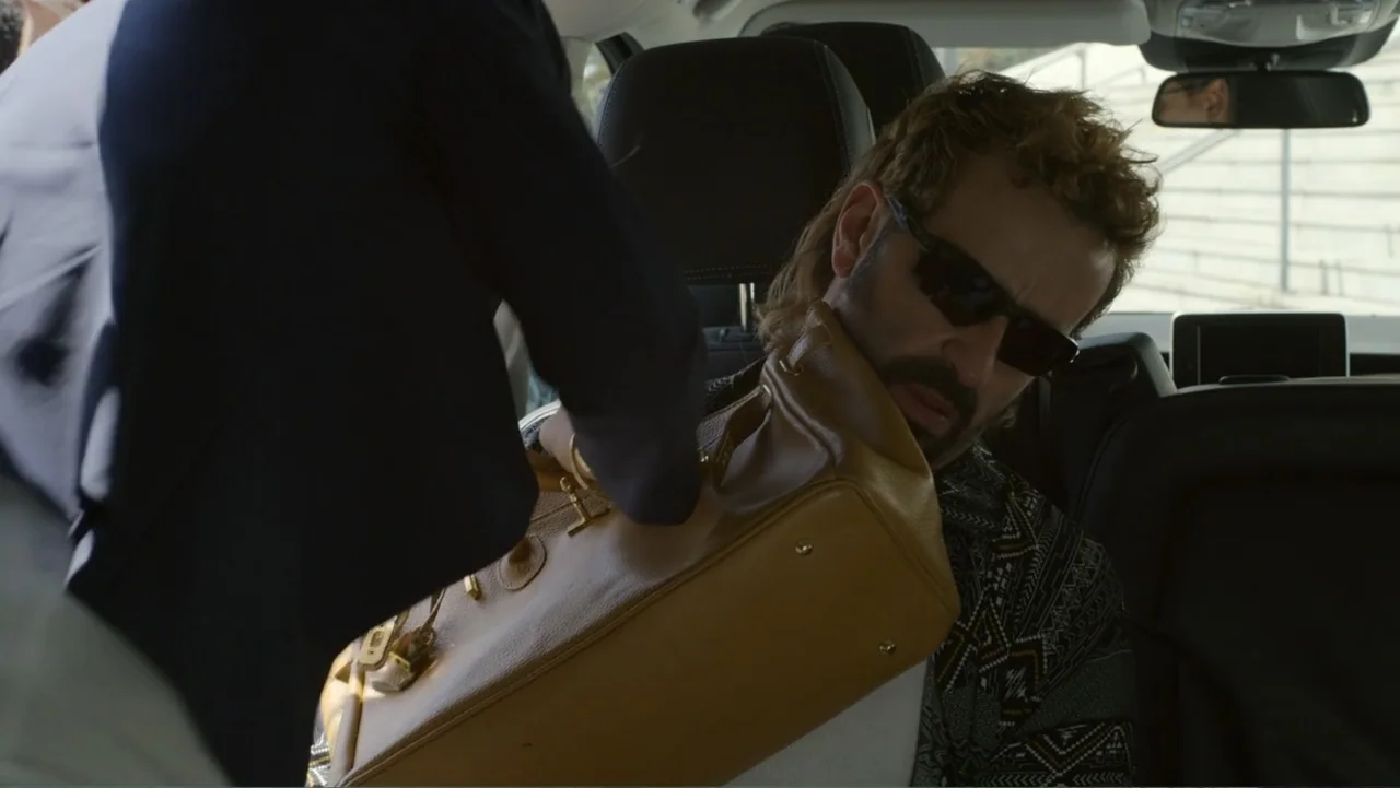 Unknown #1 movie still: medium — A dark car interior shows a passenger side seat and headrests. One individual hands a large tan leather bag to…