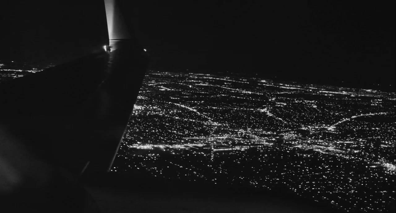 Frances Ha (2012) movie still: wide — An aerial view of an illuminated urban sprawl at night seen from high altitude. The dark silhouette of an…