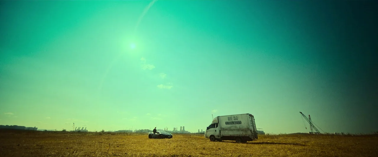 Ballerina (2023) movie still: wide — A vast, dry grassy expanse stretches toward a distant industrial skyline. A man stands beside a parked sports…