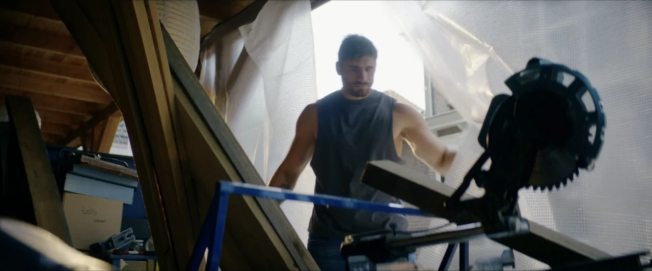 Black Lotus (2023) movie still: medium — An interior workshop space filled with lumber and construction tools under a wooden roof. A man in a…