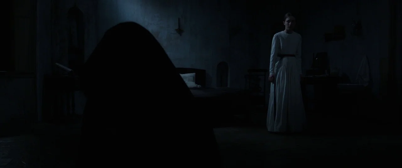 The Nun (2018) movie still: wide — A dimly lit room features stone walls, a simple wooden bed, and a single window. A woman in a white dress stands…