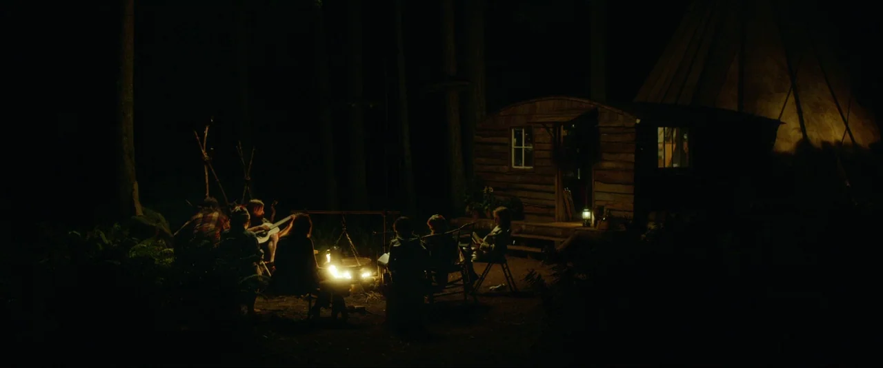 Captain Fantastic (2016) movie still: wide — A group gathers around a glowing fire in a dense forest at night. A small wooden cabin and a conical…