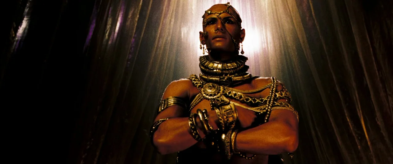 300 (2006) movie still: medium — A dark space with heavy draped fabric hangs behind an ornate, jewelry-adorned human figure. The figure stands with…