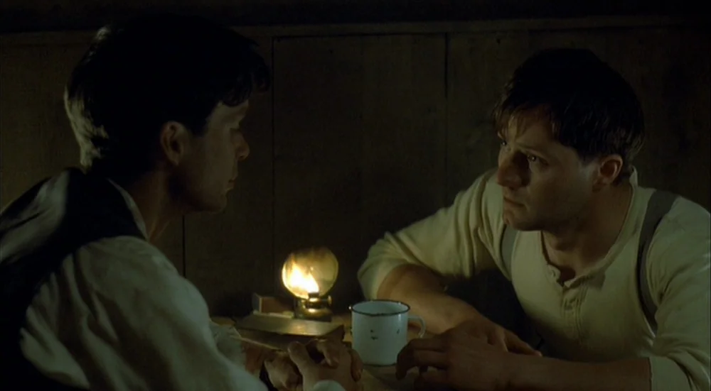 The Wind That Shakes the Barley (2006) movie still: medium — A dimly lit room with wood-paneled walls features a table between two men. The men sit…