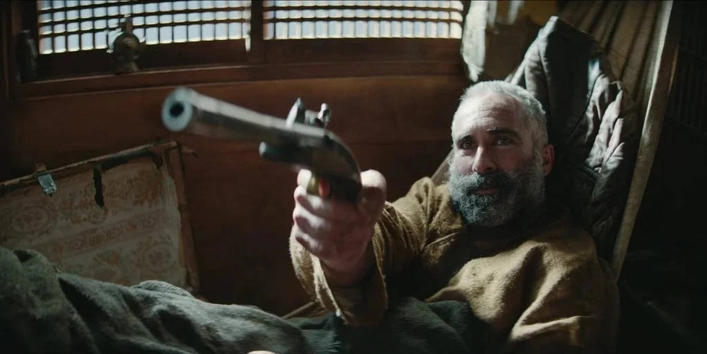 Shogun (2024) movie still: medium — A small room with wooden paneled walls and a paper-grid window. A man lying down aims a flintlock pistol…