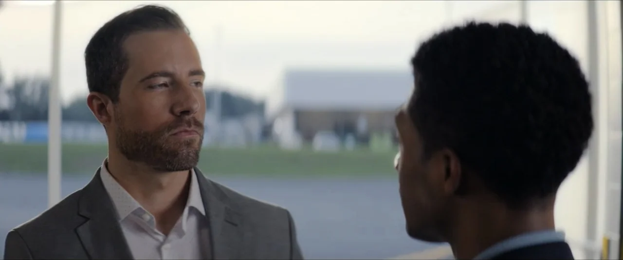 The Forge (2024) movie still: medium — Two men in business attire stand in a bright open-air structure. The men face each other while talking.