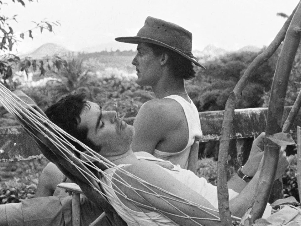 Tabu (2012) movie still: medium — Two men lounge in hammocks set up in a lush, outdoor tropical environment. One man sleeps while the other sits…