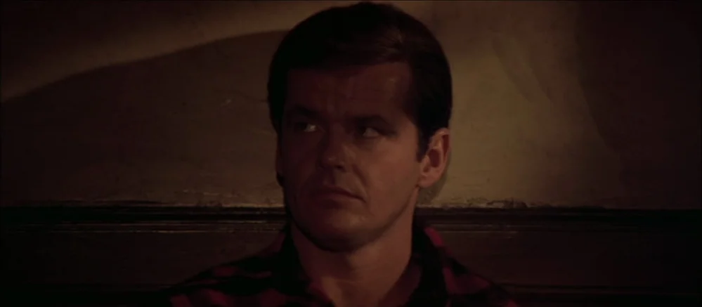 Carnal Knowledge (1971) movie still: closeup — A dimly lit indoor space with plain wall texture behind the central subject. A man in a red and black…