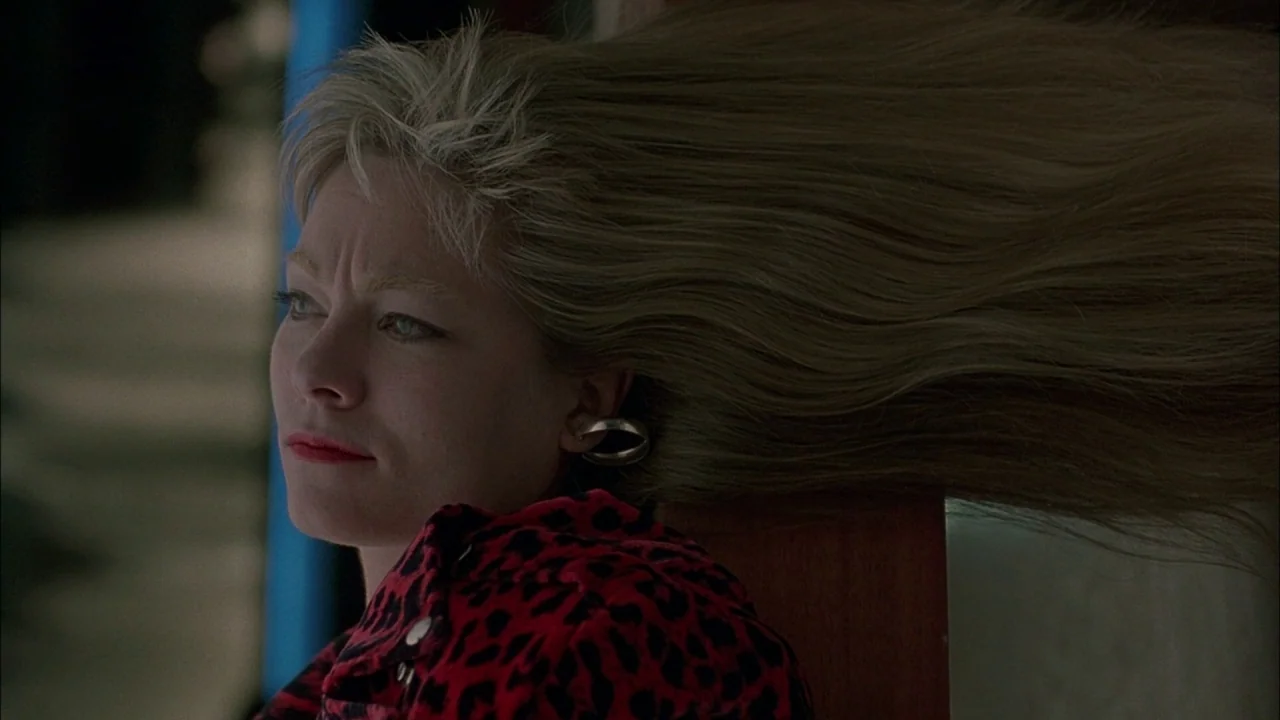 Whore (1991) movie still: closeup — A woman stands on a porch with wooden elements and a blue pillar. Her long blonde hair is blown forcefully to…