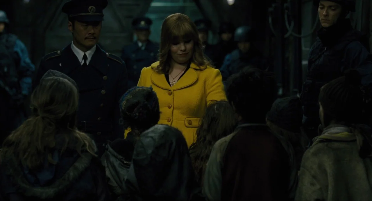 Snowpiercer (2013) movie still: medium — A dimly lit industrial hallway features a woman in a yellow jacket facing children. Uniformed men stand…