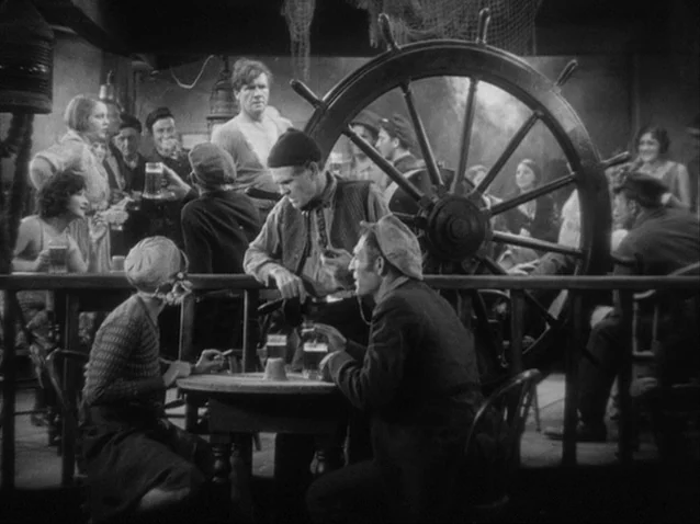 The Docks of New York (1928) movie still: medium — A crowded maritime-themed pub interior features a large wooden ship wheel as decor. People in…