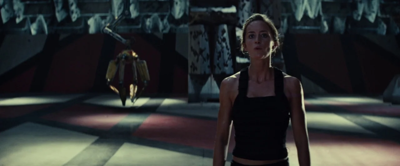 Edge of Tomorrow (2014) movie still: medium — A dark industrial hangar features a geometric red and white tiled floor pattern. A lone woman stands…
