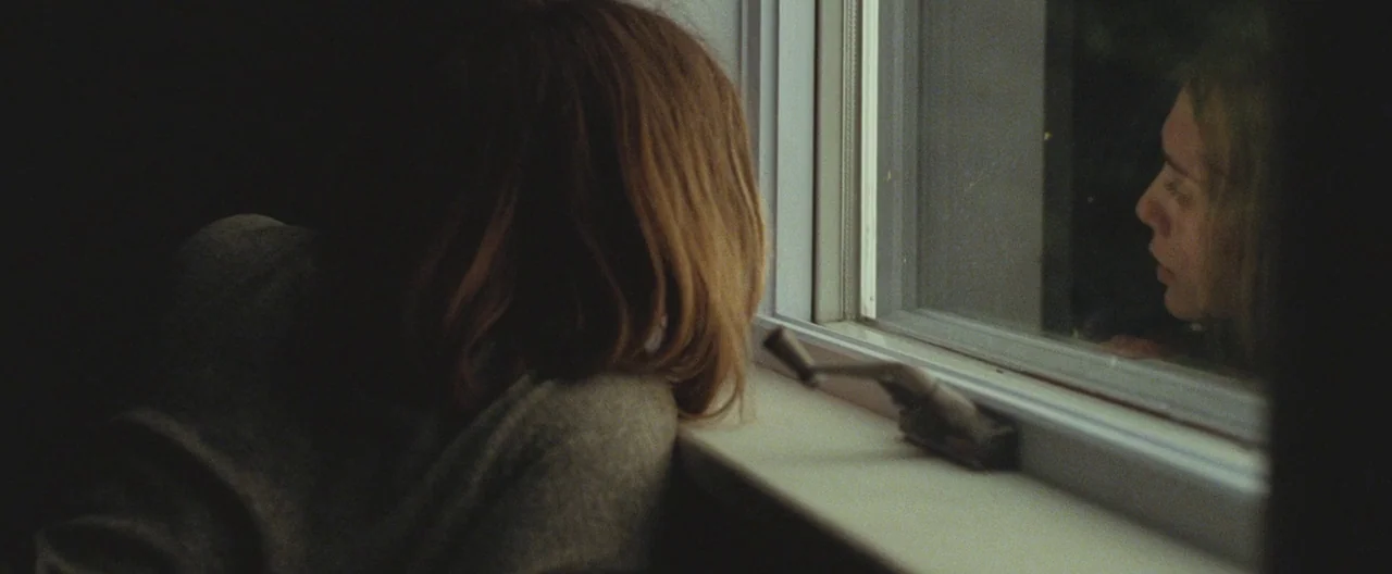 Martha Marcy May Marlene (2011) movie still: medium — A woman sits indoors looking through a window at night. Her reflection is visible on the glass…