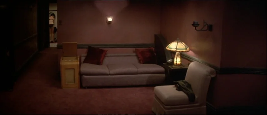 Blue Velvet (1986) movie still: wide — A dimly lit hotel seating area features deep red walls and carpeting. A sofa with dark pillows and a…