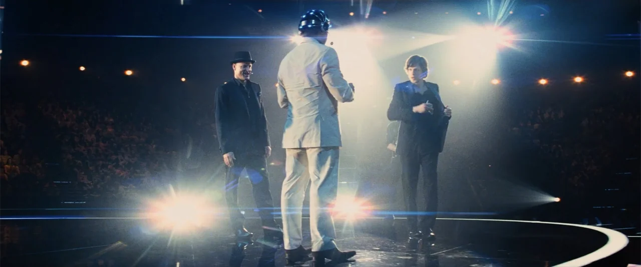 Now You See Me (2013) movie still: fullbody — A dark, expansive stage is lit by intense spotlights from behind the subjects. Three men stand on the…