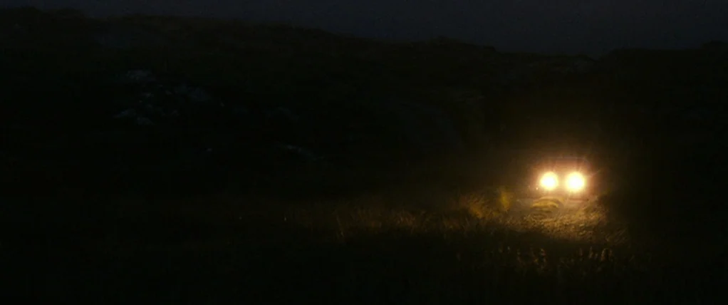 Ondine (2009) movie still: wide — A dark, uneven field at night is partially visible under a thin layer of night air. Two bright headlights from an…