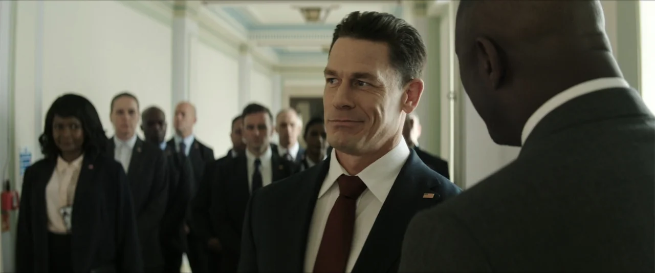 Heads of State (2025) movie still: medium — A long, sterile government corridor with white walls and a large group of agents. John Cena stands in…