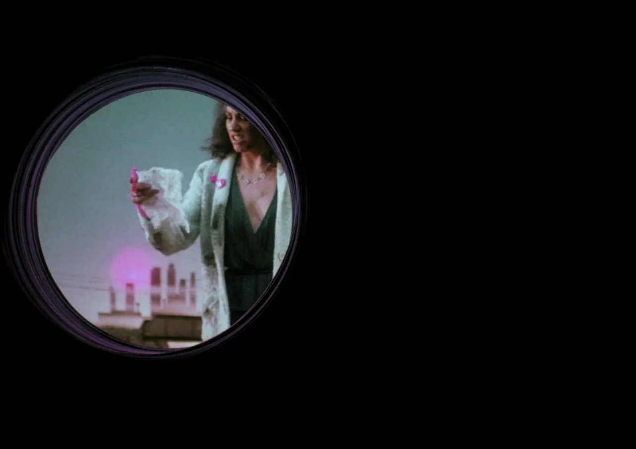 Human Highway (1982) movie still: medium — A woman stands on a rooftop against a dark sky at night. She is viewed through a circular aperture as she…