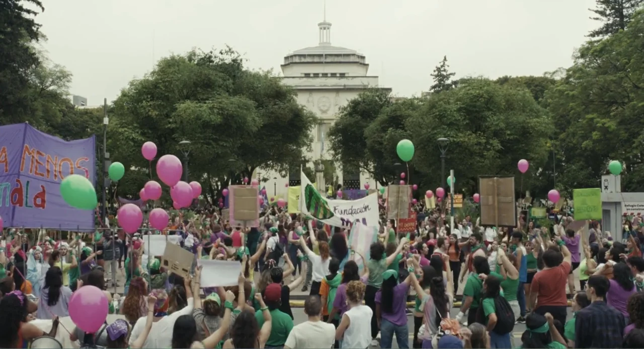 Belén (2025) movie still: wide — A public plaza sits before a large neoclassical government building under an overcast sky. A large crowd gathers in…