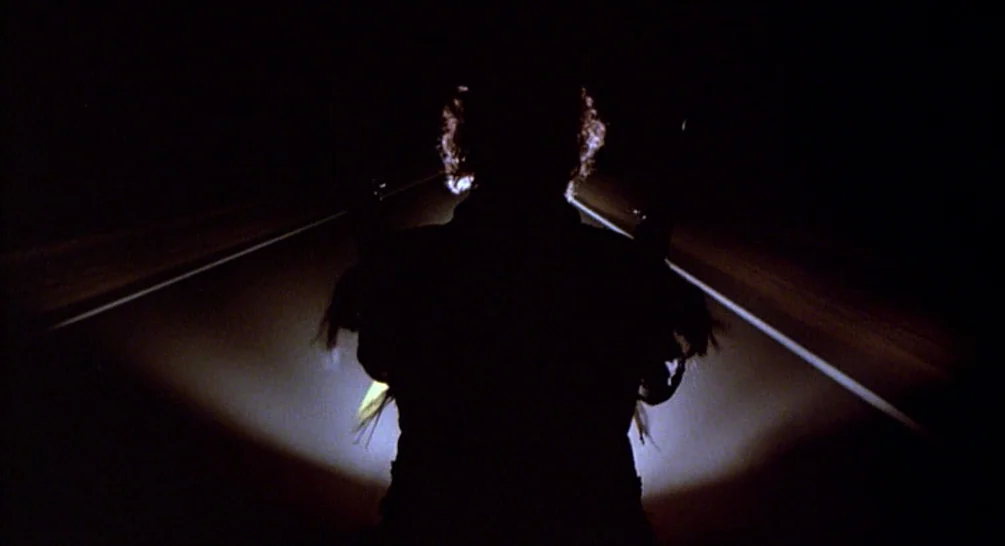 Raising Arizona (1987) movie still: medium — A dark, empty paved road at night with two lane markings visible. A female figure stands in the center…