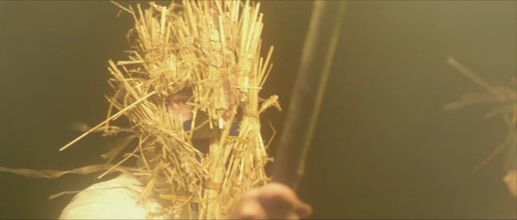 Kill List movie still: closeup — A figure wears a crude mask constructed from bundled straw in a hazy environment. The person grips a wooden shaft…