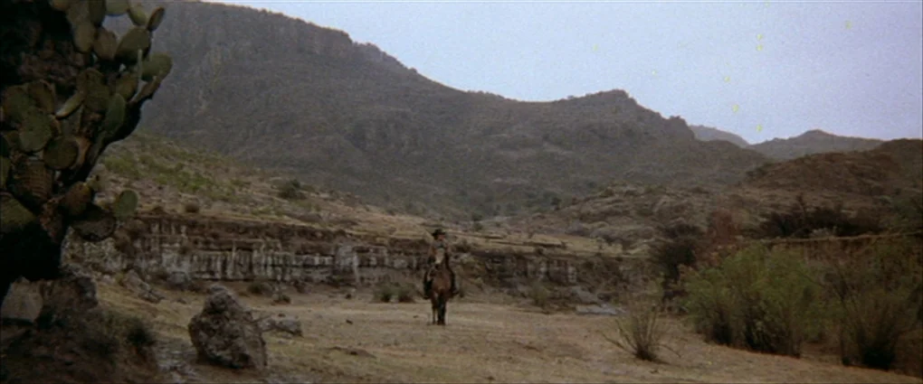 Pat Garrett & Billy The Kid (1973) movie still: wide — A vast arid landscape features low rocky cliffs and sparse dry vegetation in Mexico. A lone…