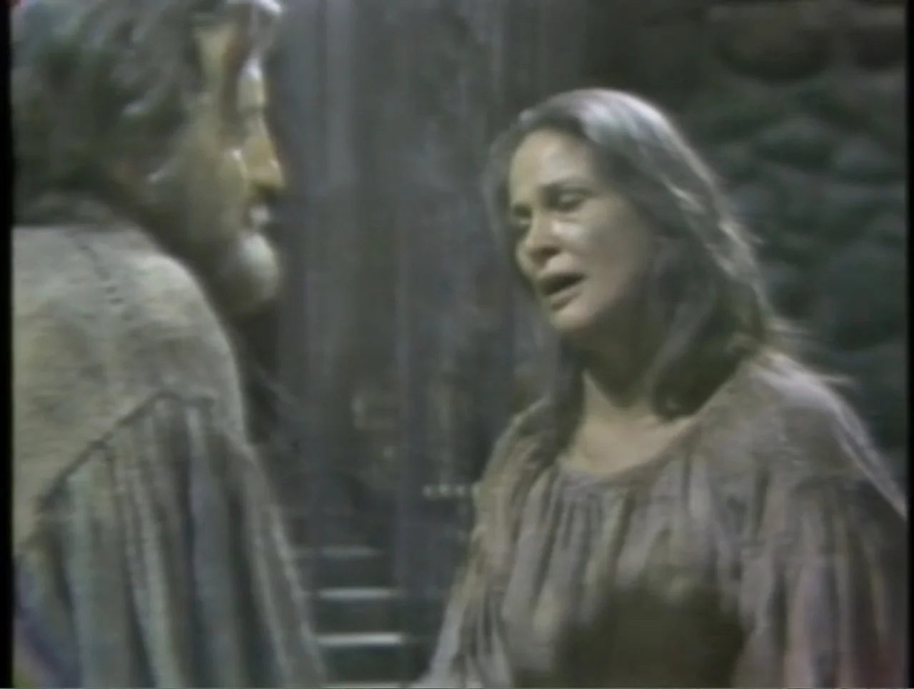 The Crucible (1996) movie still: medium — A dimly lit stone chamber with rough masonry walls features a man and woman. A middle-aged woman in a…