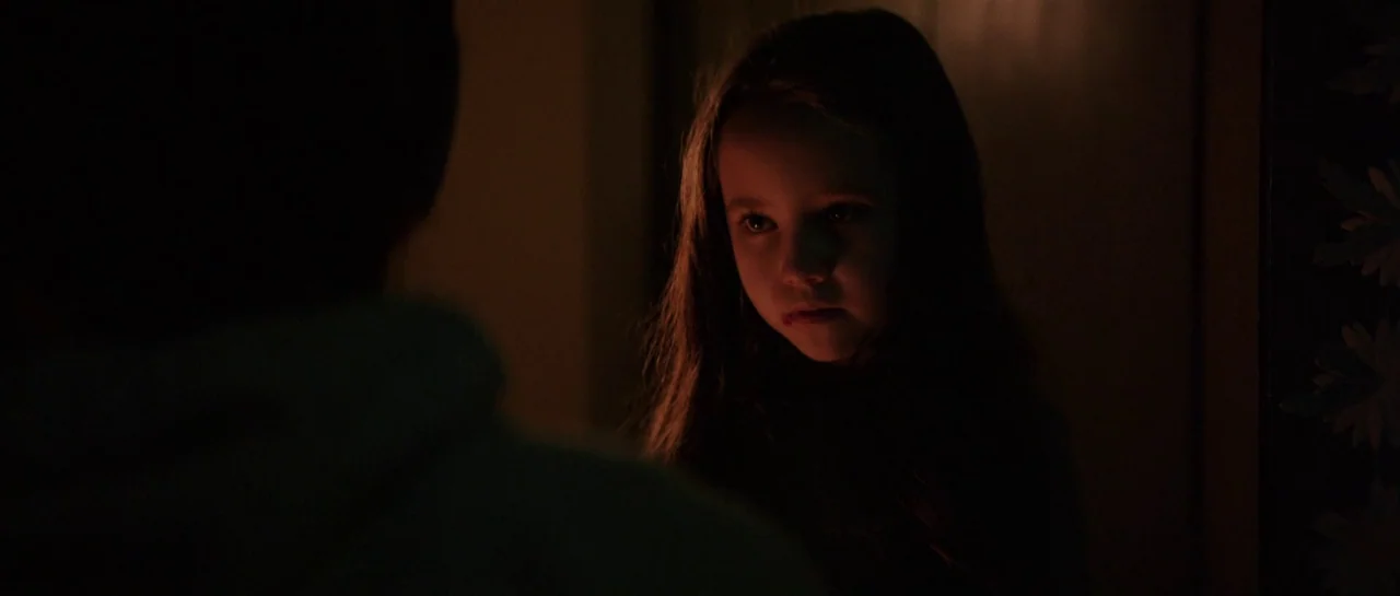 The Boogeyman (2023) movie still: closeup — A dimly lit room with heavy shadows obscuring the background and sides of the frame. A young girl is…