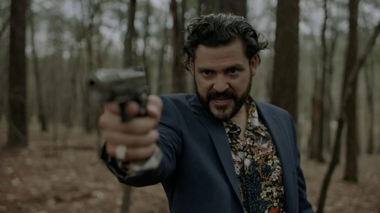 Die Hart (2023) movie still: medium — A man stands in a wooded area with tall trees in the soft-focused background. He holds a black handgun pointed…
