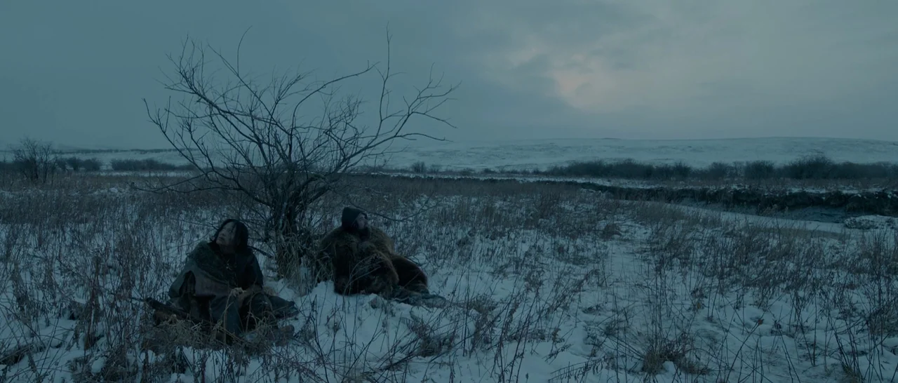 The Revenant (2015) movie still: wide — A snowy, barren tundra landscape with a small, leafless tree in the center. Two men in heavy, worn winter…