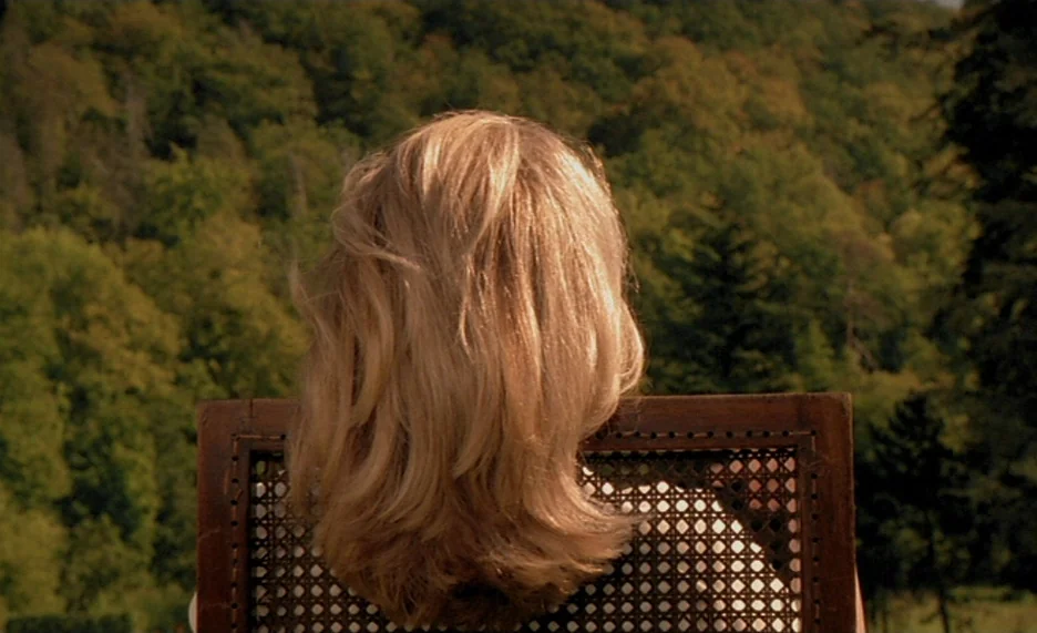 Pola X (1999) movie still: closeup — A woman sits in a wooden cane-back chair outdoors in front of a dense forest. The camera is positioned behind…