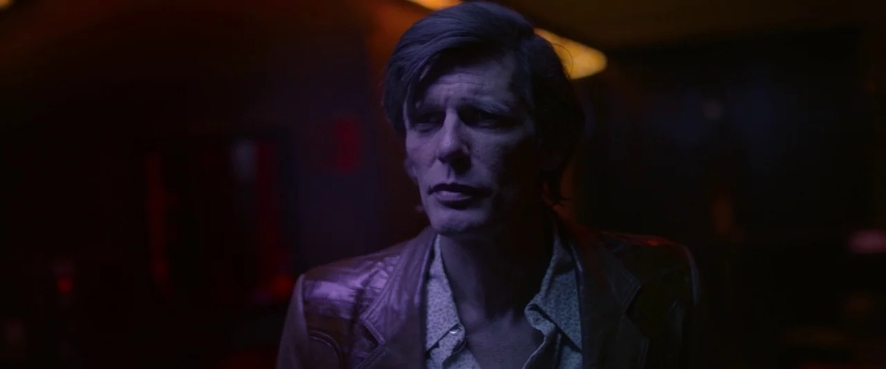 Blaze (2018) movie still: medium — A dark, dimly lit lounge space filled with deep purple shadows and saturated color. A middle-aged man stands in…