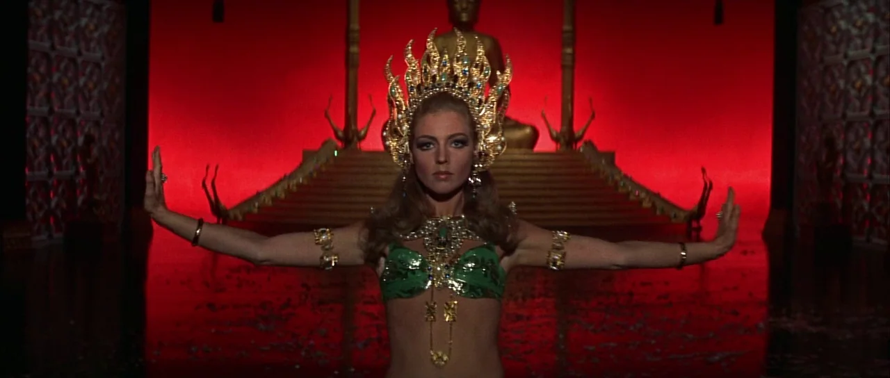 Casino Royale (1967) (1967) movie still: medium — A dark, red-lit temple interior features a central golden buddha statue on steps. A woman stands…