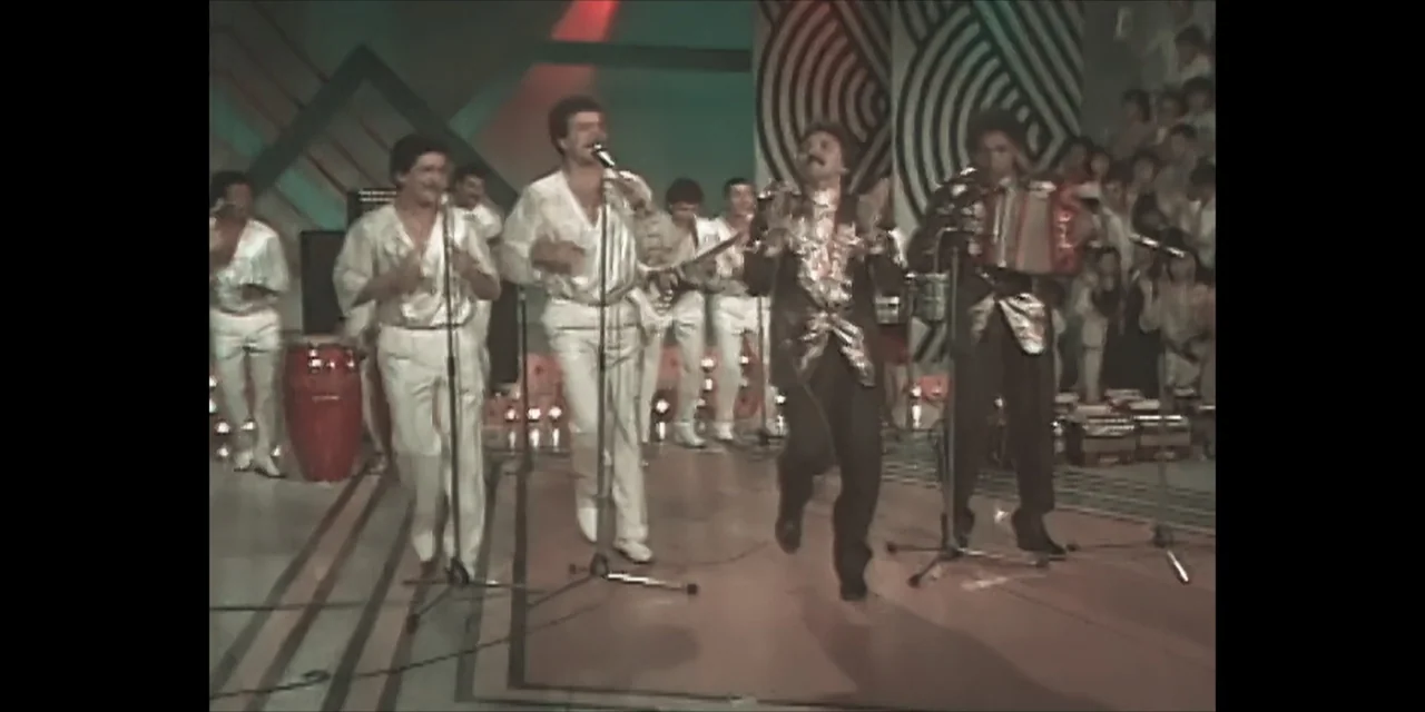 Leyenda Viva (2022) movie still: fullbody — A 1980s television studio stage features geometric background patterns and microphones on stands. Four…