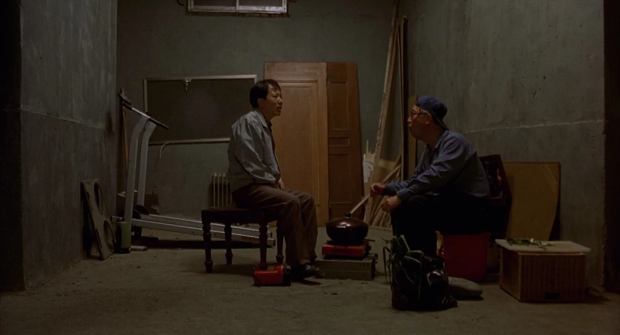 Barking Dogs Never Bite (2000) movie still: fullbody — A sparse, concrete basement room contains scattered furniture and domestic items on the…