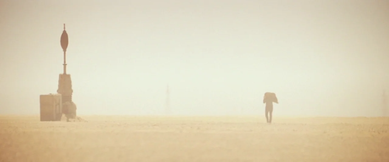 Star Wars: The Force Awakens (2015) movie still: wide — A vast, hazy desert plain under a bright, featureless sky stretches to the horizon. A lone…