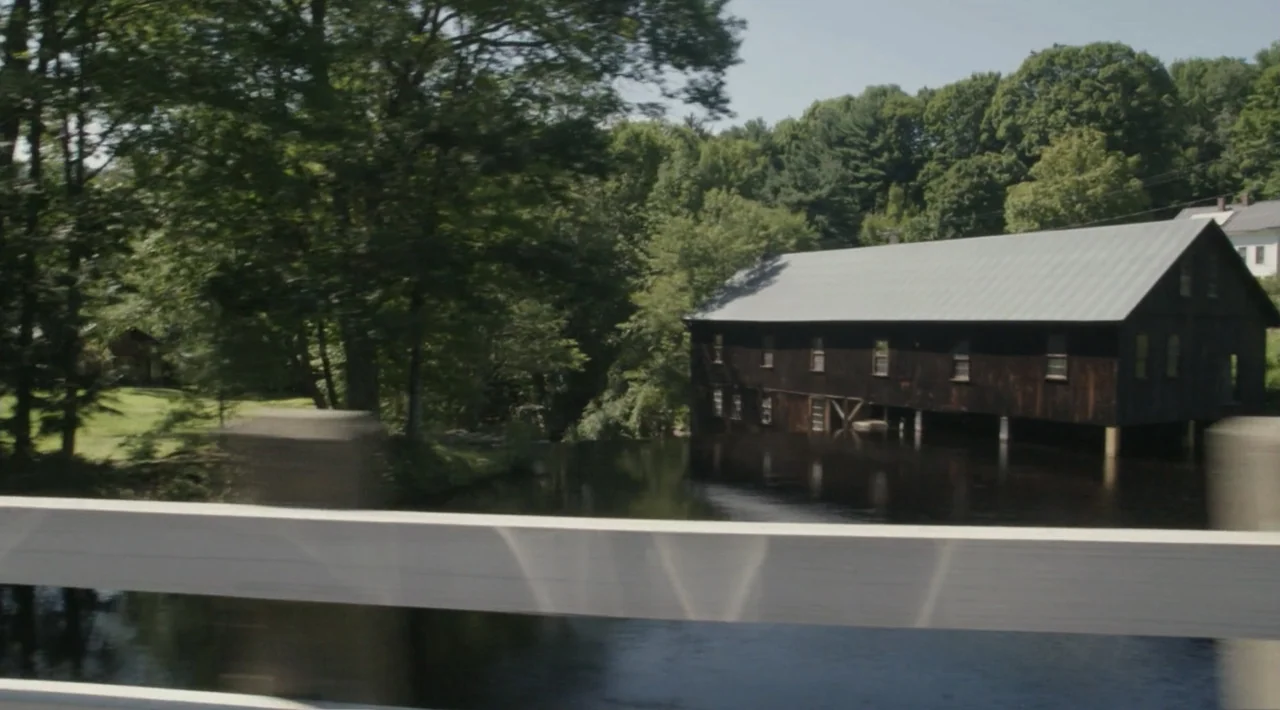 The Divided Soul of America (2019) movie still: establishing — A dark wooden mill stands on stilts over a calm river in a forest. Lush green trees…