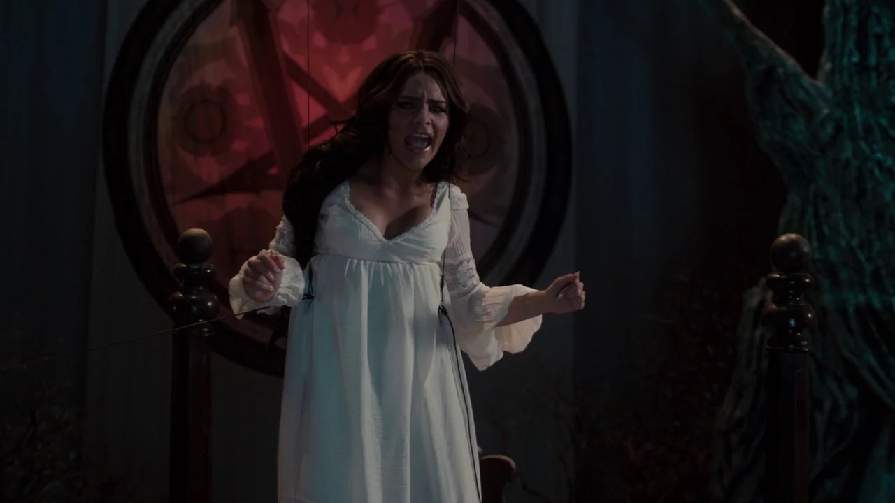 Fright Night (2011) movie still: medium — A woman stands on a stage in a white dress with puppet strings attached. A large, dark circular clock-like…