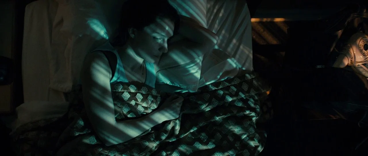 The Imposter (2012) movie still: medium — A dark bedroom at night with patterned bedding and white pillows. A woman lies under the blankets with…