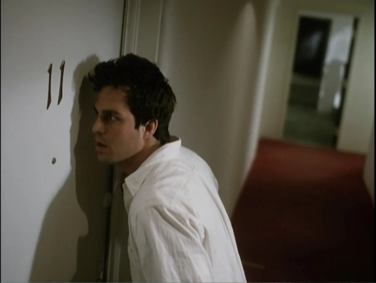 Apartment 12 (2001) movie still: medium — A dimly lit hotel hallway features a carpeted floor and a plain door. A man in a white shirt leans forward…