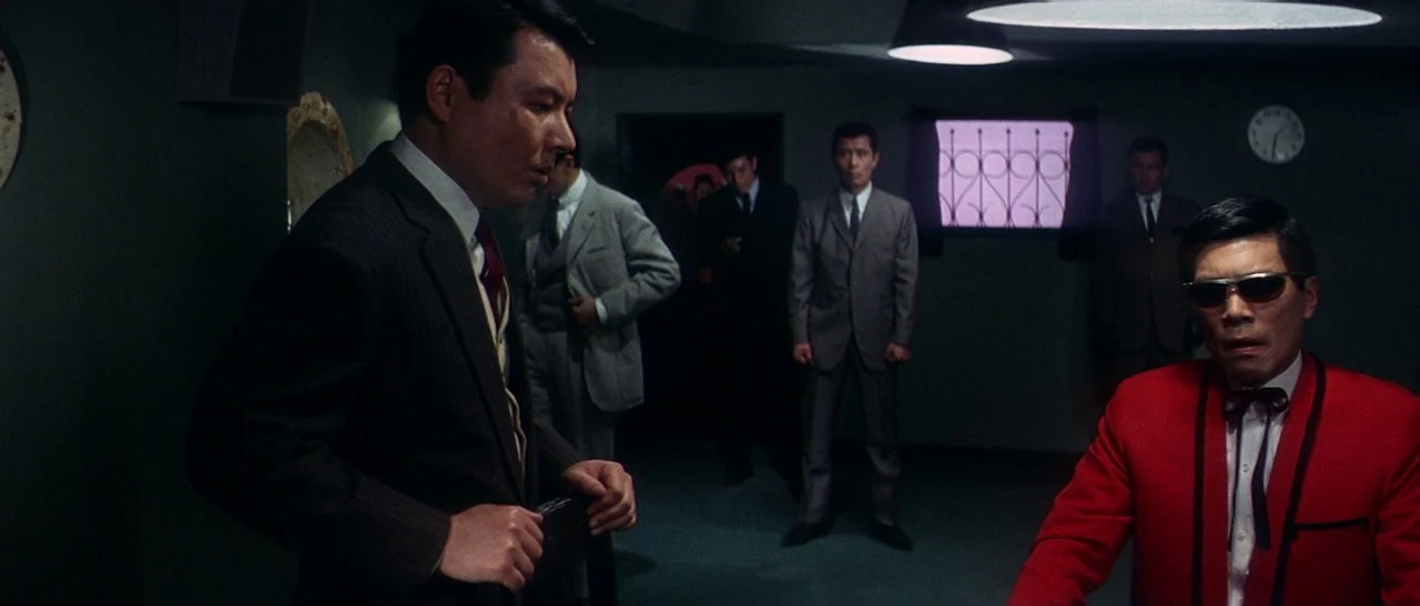 Tokyo Drifter (1966) movie still: medium — A dark, dimly lit lounge features green walls and a wall-mounted clock. Several men in business suits…